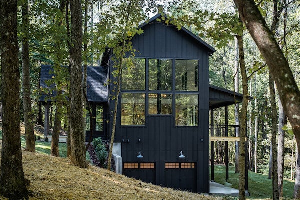 A black modern cabin with large windows and black garage doors in a wooded area.