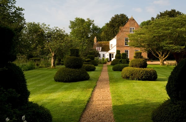 An white English cottage in the European countryside with a large red brick gable, sitting on a lawn with green trees and sculpted bushes. 