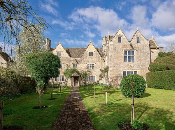 A large manor house on a green lawn with a weathered stone exterior, dilapidated windows, and several high-pitched gables.