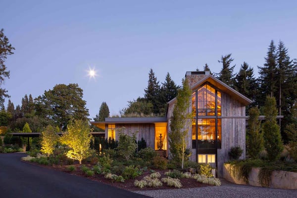 Modern barndominium house with a big window design sitting among trees as the sun sets in the background.