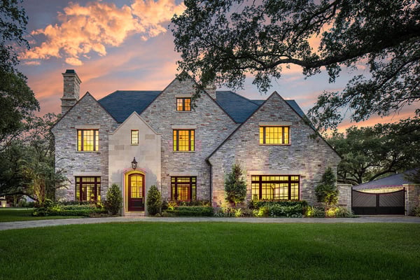 A modern manor style house with a stone facade, black roof, and black grid windows in front of a sunset and behind a green well-kept lawn.