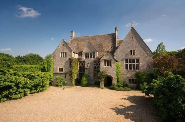 A medieval manor house with antiquated windows, old stone, and a sunken-in roof surrounded by green trees and bushes.