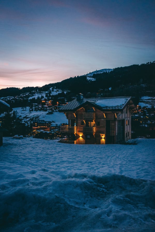 Mountain farm house set in an open area at the foot of a hill. As the sun begins to set the house’s exterior lights are switched on accenting the beautiful roofline and attributes of the home.