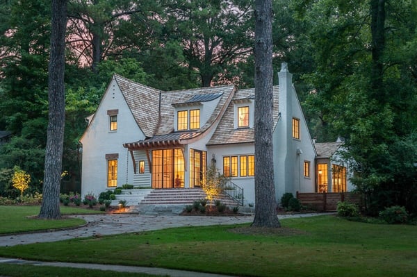 A white transitional tudor style home with a curved roof encircled by tall green trees