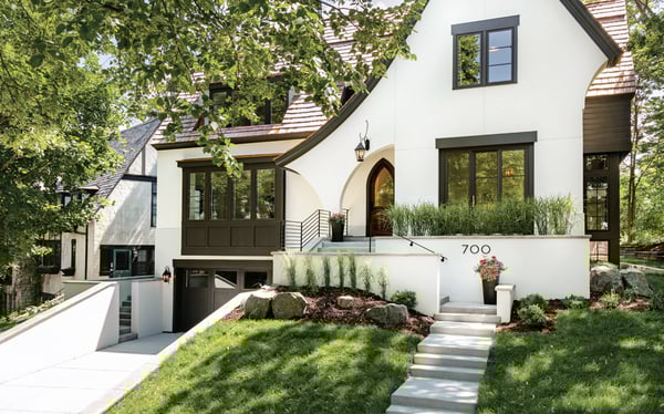  A modern manor tudor house with a white exterior, large glass windows with black trim, and a central arched doorway. 