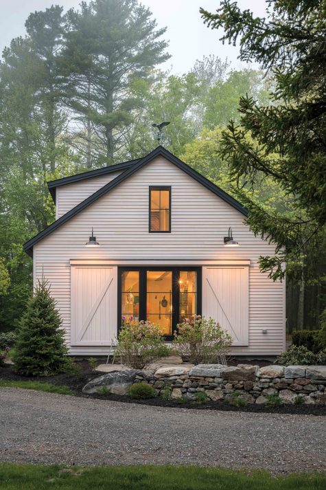 A small, cozy white modern barndominium style house built amongst trees with a rock wall in front of a big window.